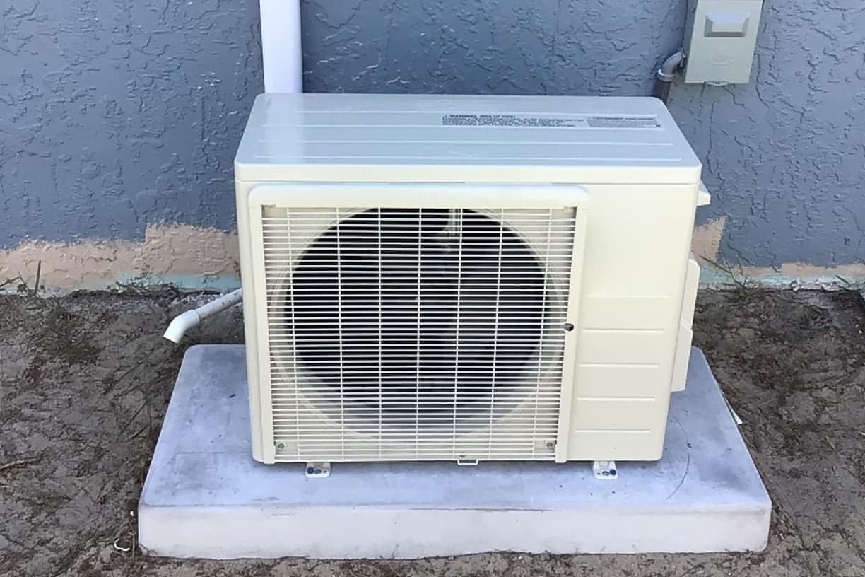 A ductless mini-split outdoor unit on a pad against a blue wall.