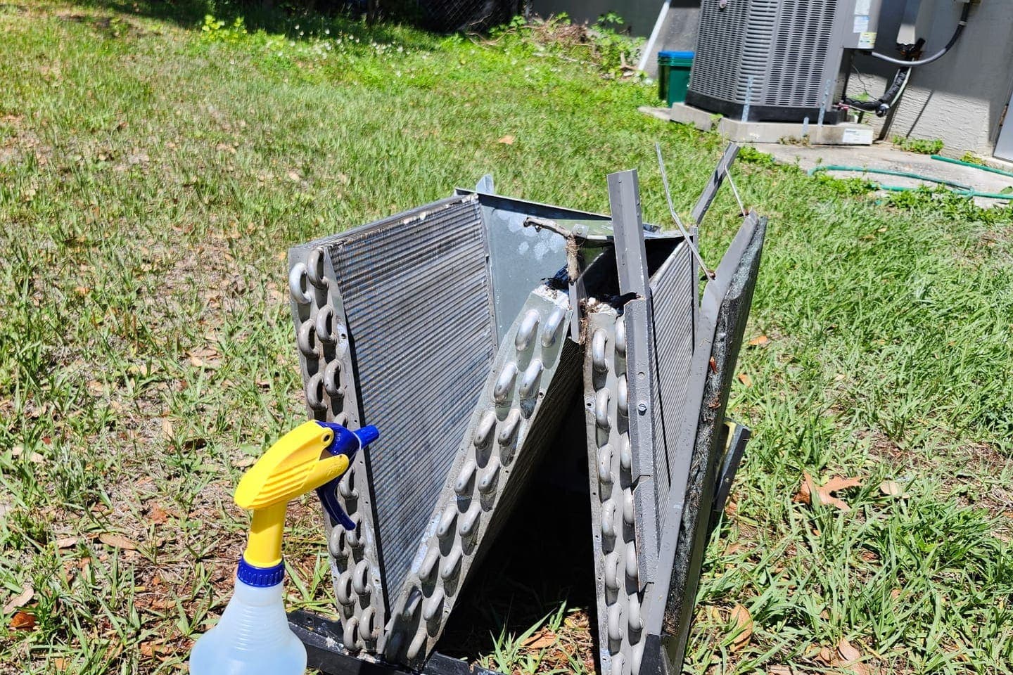 An evaporator coil lifted out and laid on the grass beside a coil-cleaner spray bottle.