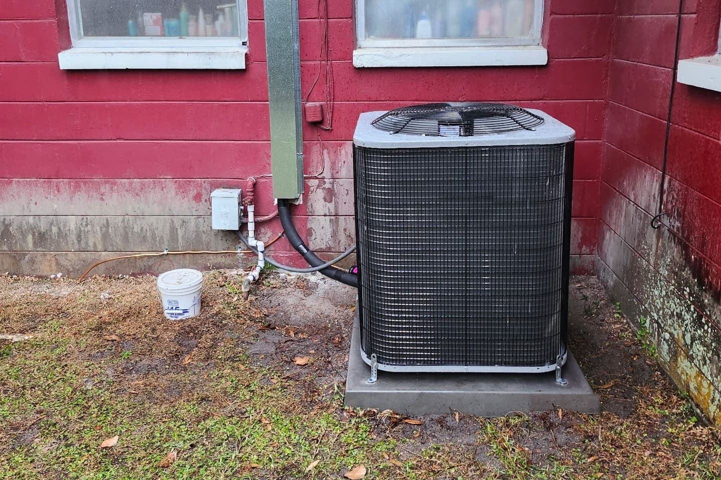 An outdoor condenser standing on a pad against a red-painted wall.