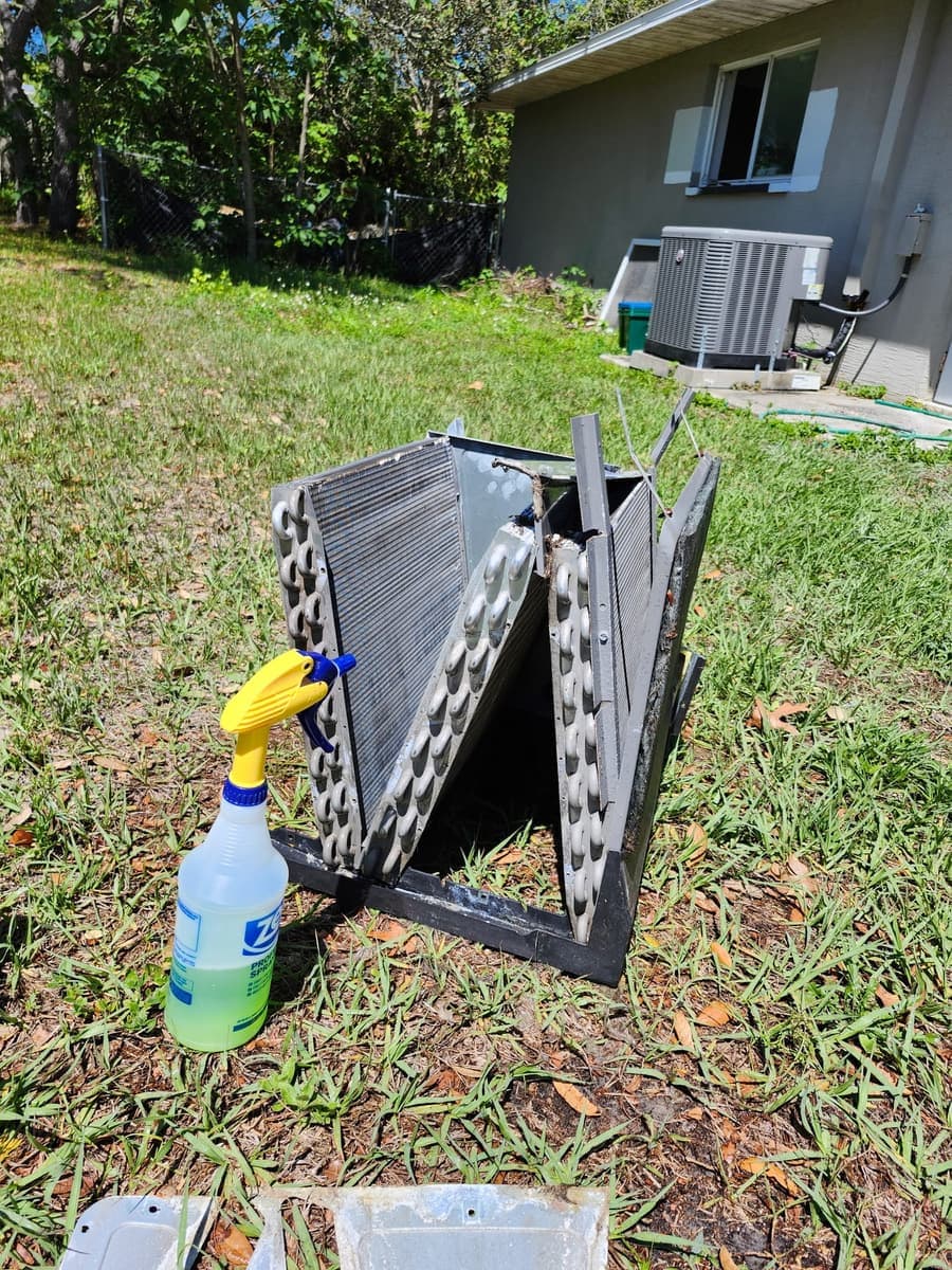 An evaporator coil lifted out and laid on the grass beside a coil-cleaner spray bottle.