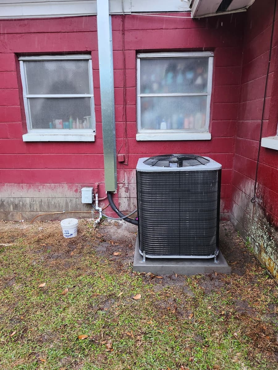 An outdoor condenser standing on a pad against a red-painted wall.