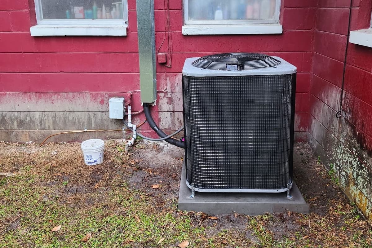 An outdoor condenser standing on a pad against a red-painted wall.