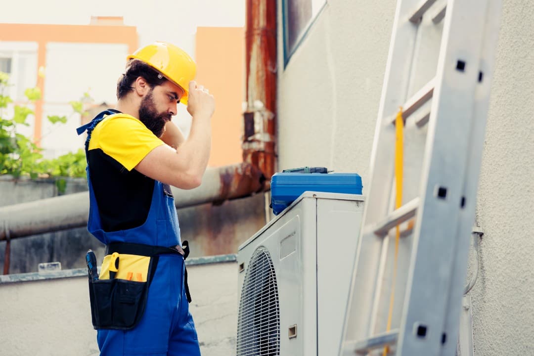 Technician in a hard hat and tool belt beside an outdoor mini-split unit and a ladder
