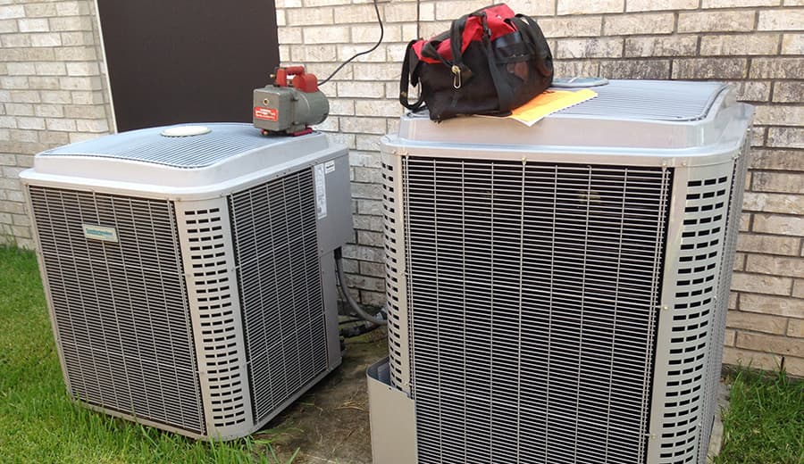 Old and new outdoor AC condensers side by side next to a brick home during a system replacement
