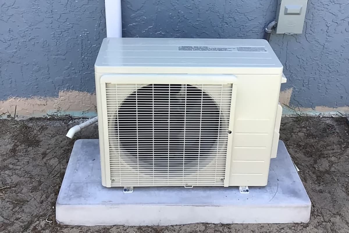 A ductless mini-split outdoor unit on a pad against a blue wall.