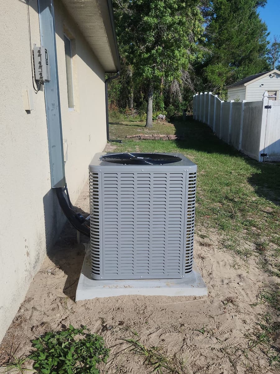 A gray outdoor condenser on a sand pad beside a house wall.
