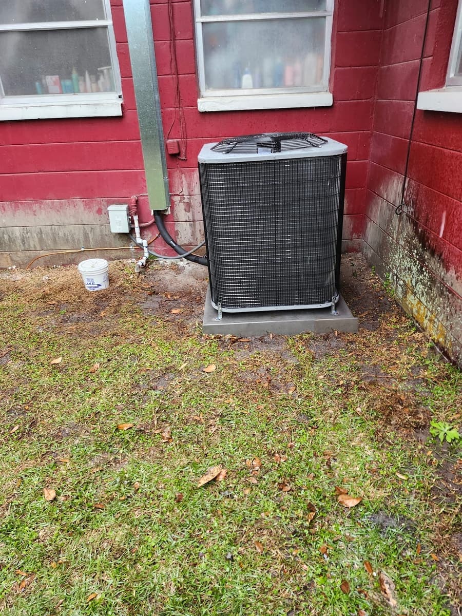 An outdoor condenser unit on a pad beside a red wall and electrical disconnect.