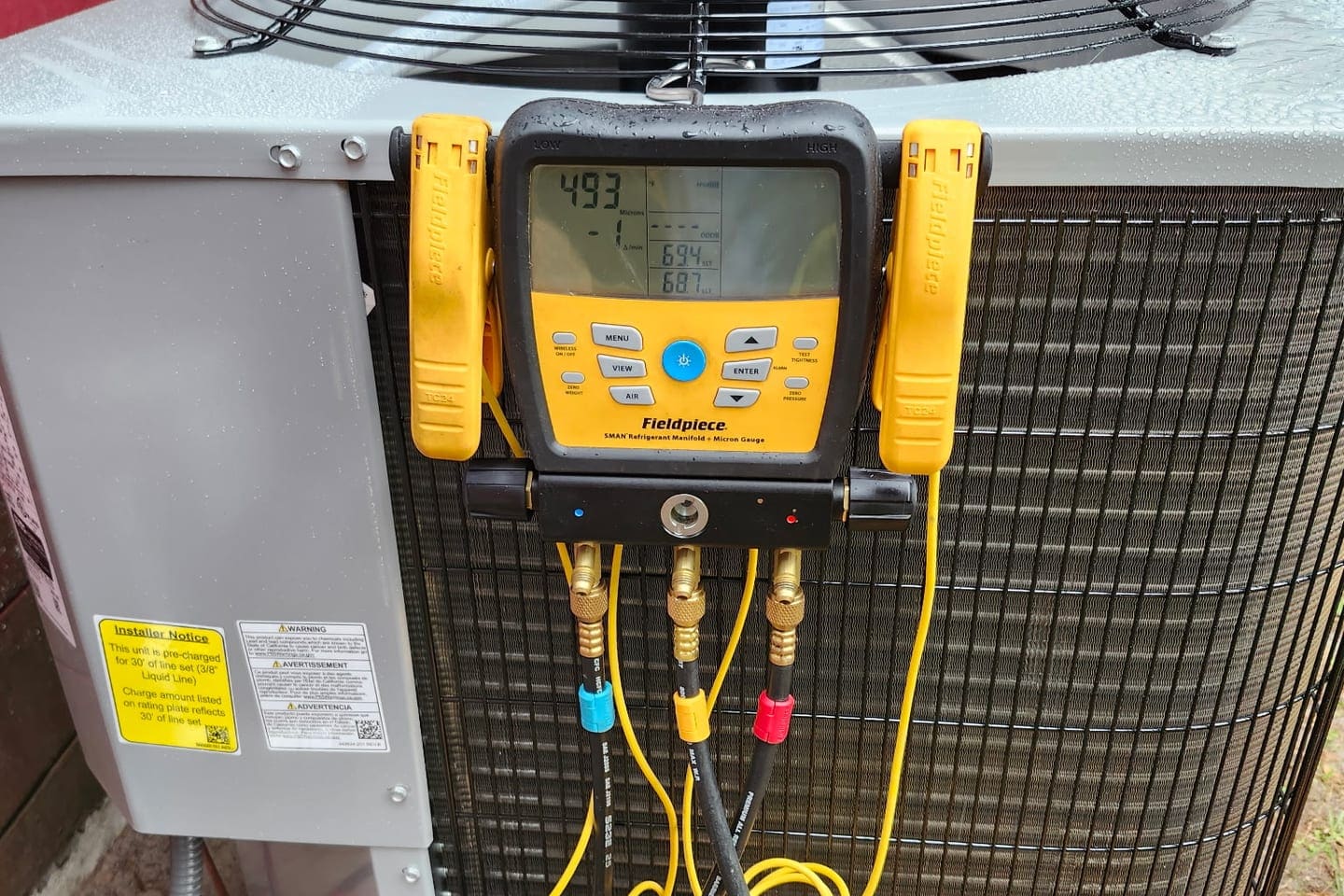 A digital gauge manifold with yellow hoses clamped to a condenser.