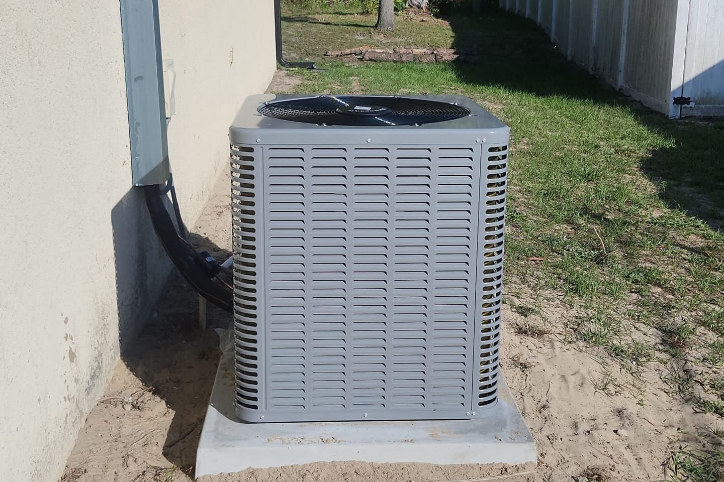 A gray outdoor condenser on a sand pad beside a house wall.