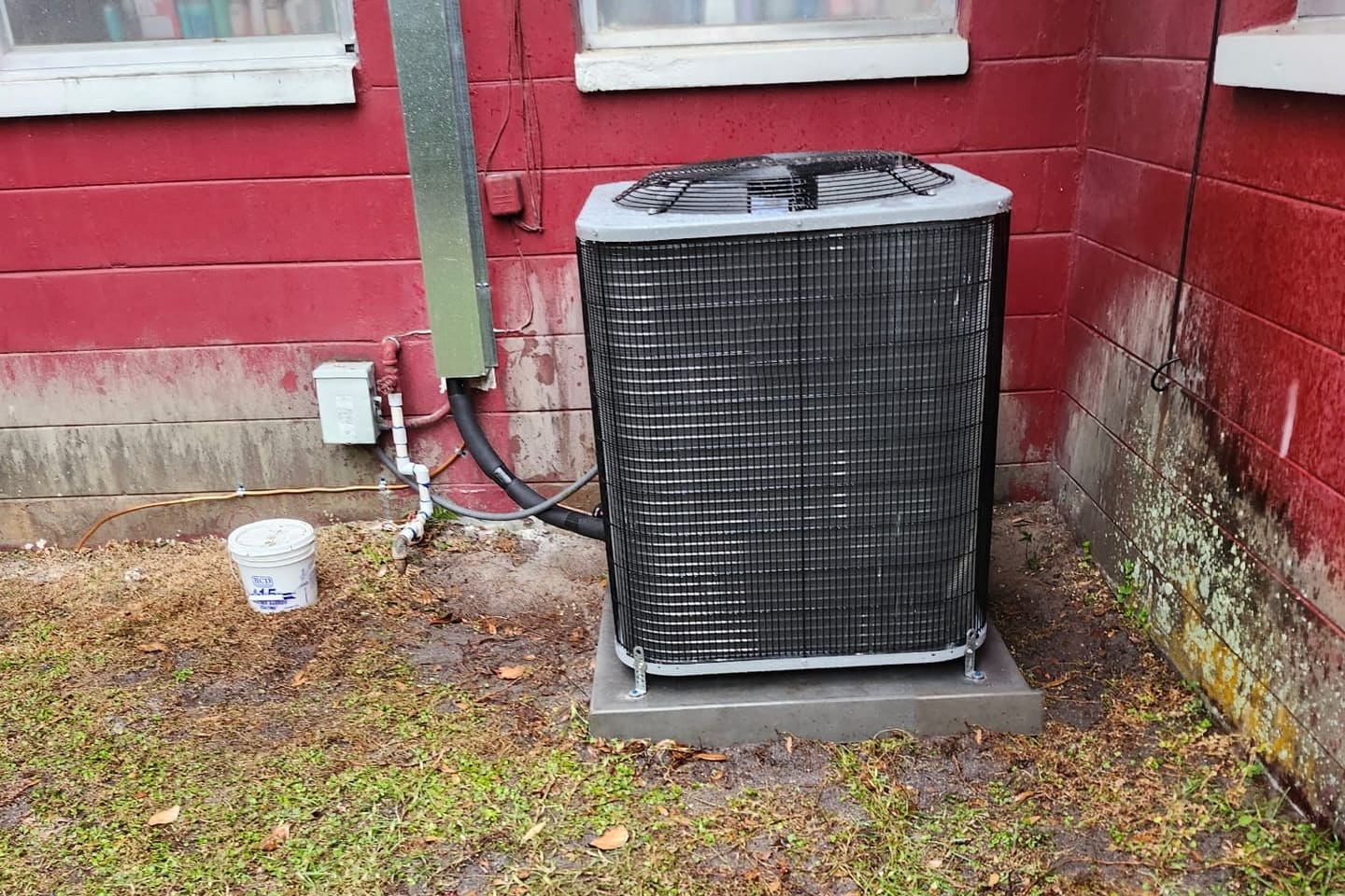 An outdoor condenser unit on a pad beside a red wall and electrical disconnect.